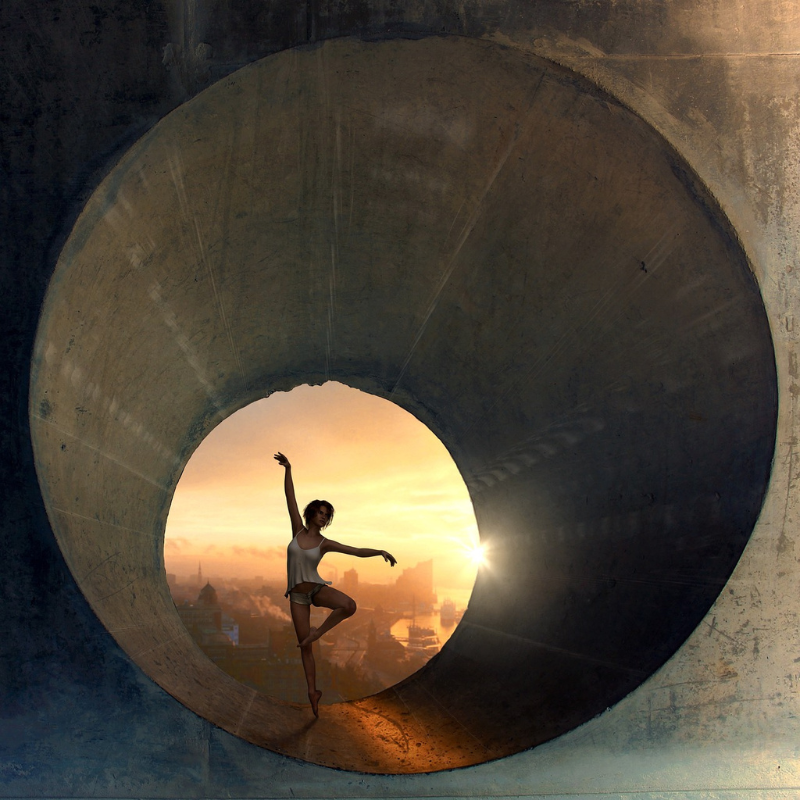 Une personne se tient debout sur un pied dans une pose de ballet à l’intérieur d’une grande structure circulaire en béton. Le coucher de soleil crée une toile de fond avec un ciel orange et un paysage urbain au loin, comme pour vaincre la fatigue avec grâce et détermination.