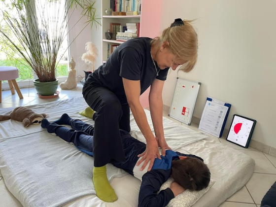 Un enfant vêtu de vêtements sombres reçoit un massage du dos alors qu'il est allongée face contre terre sur un tapis dans une pièce bien éclairée. Sophie Husson exerce une pression avec ses mains sur le dos.