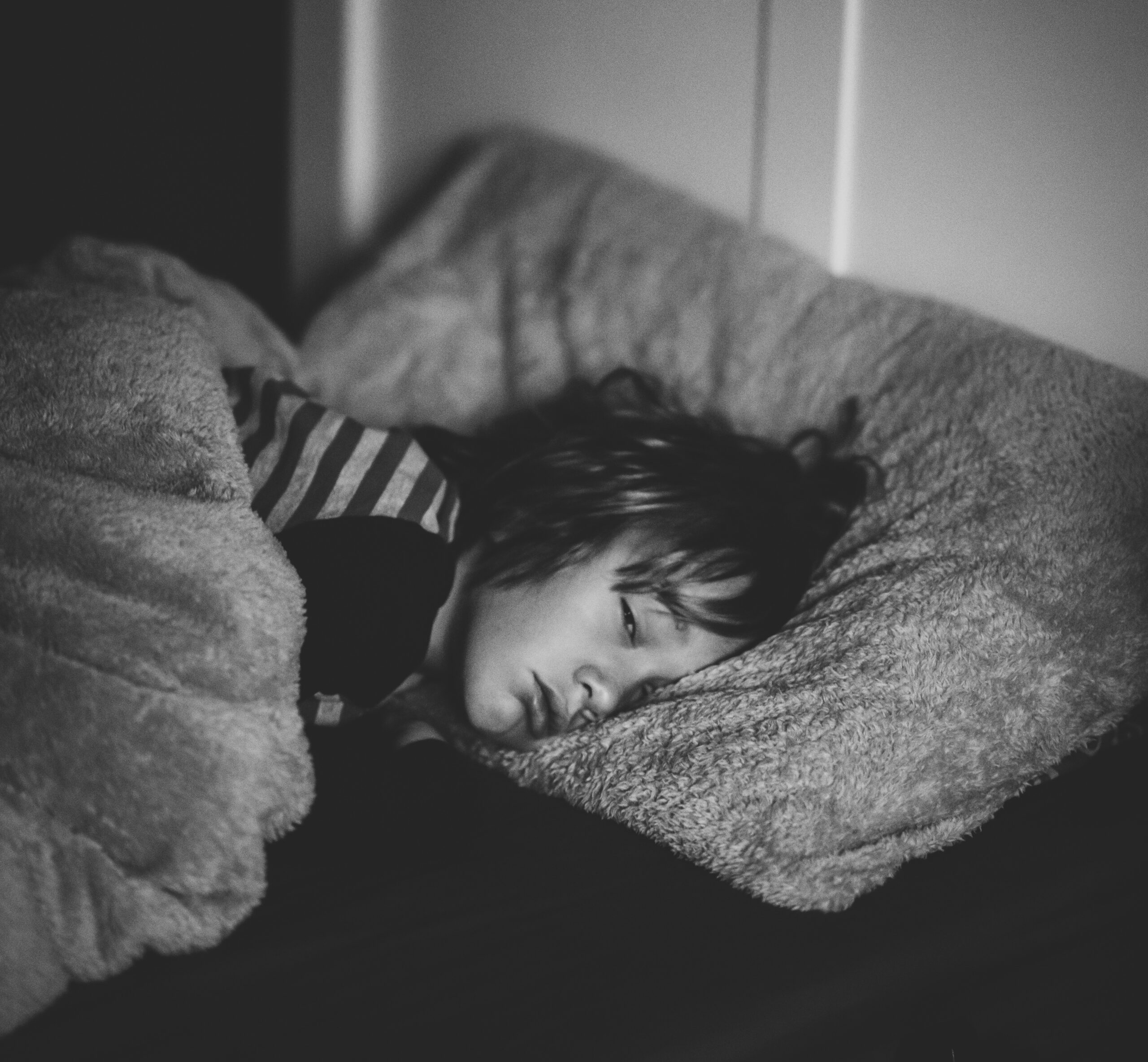 Un enfant aux yeux fermés est allongé sur un lit, recouvert d’une couverture et posant sa tête sur un oreiller, dans une pièce faiblement éclairée, cherchant peut-être du réconfort à ses troubles du sommeil chez l’enfant lié à l’hyperactivité.