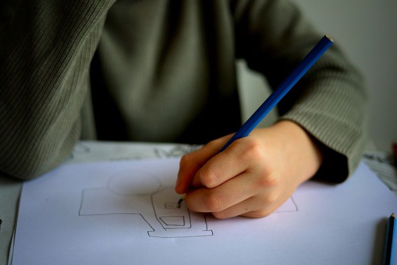 Un enfant portant une chemise à manches longues dessine sur une feuille de papier avec un crayon bleu, peut-être sujet à des problèmes de sommeil chez l’enfant lié à l’hyperactivité.