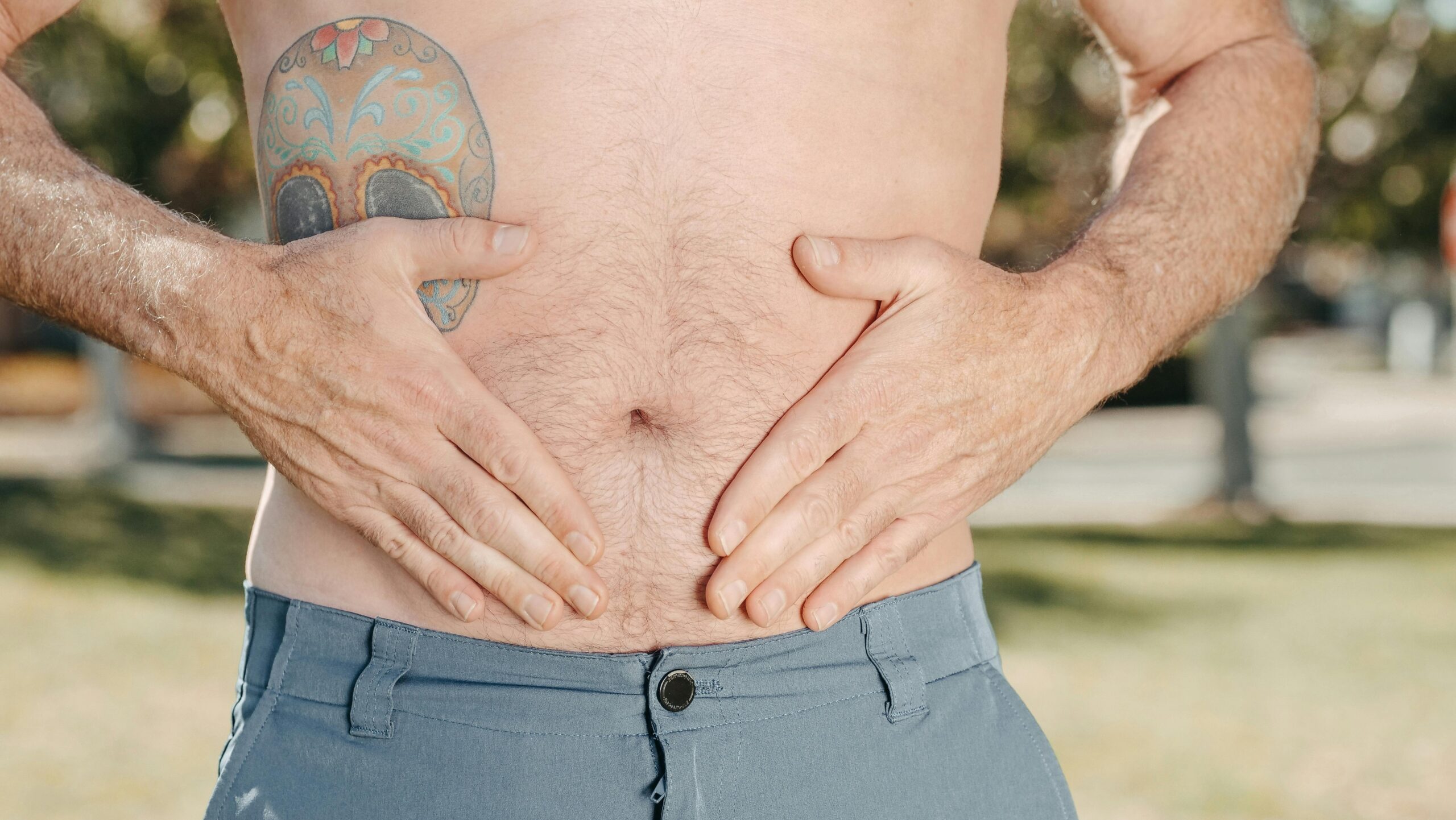 Une personne torse nu avec un tatouage sur l'épaule se tient le ventre à deux mains, debout à l'extérieur.